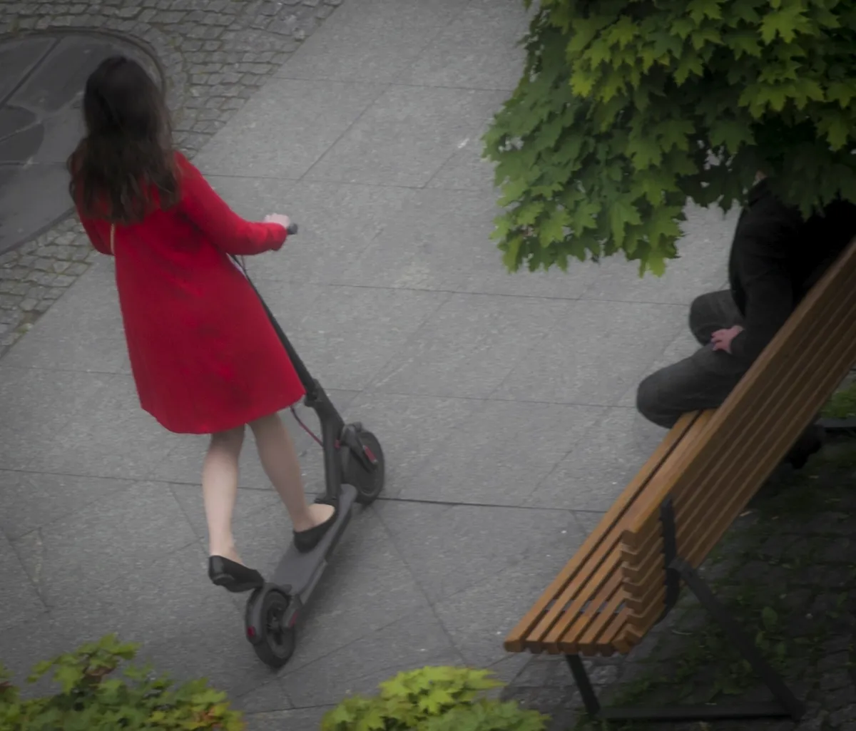 Woman in a red dress riding an electric scooter past a bench seen from above